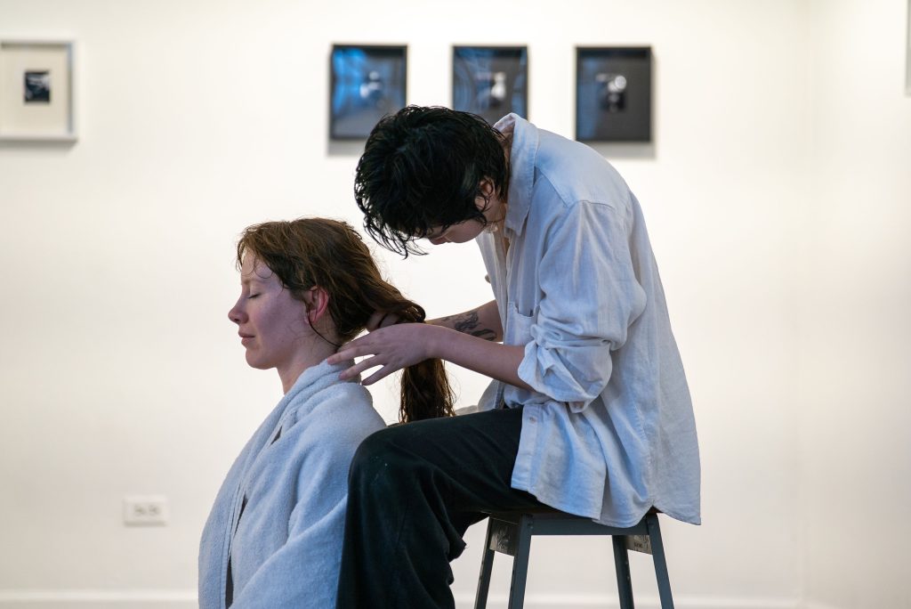 A person with short black hair and a white button-up shirt sits on a stool behind a red-haired woman, who sits on the ground wrapped up in a towel with eyes closed. The person behind her gathers her red hair together. 
