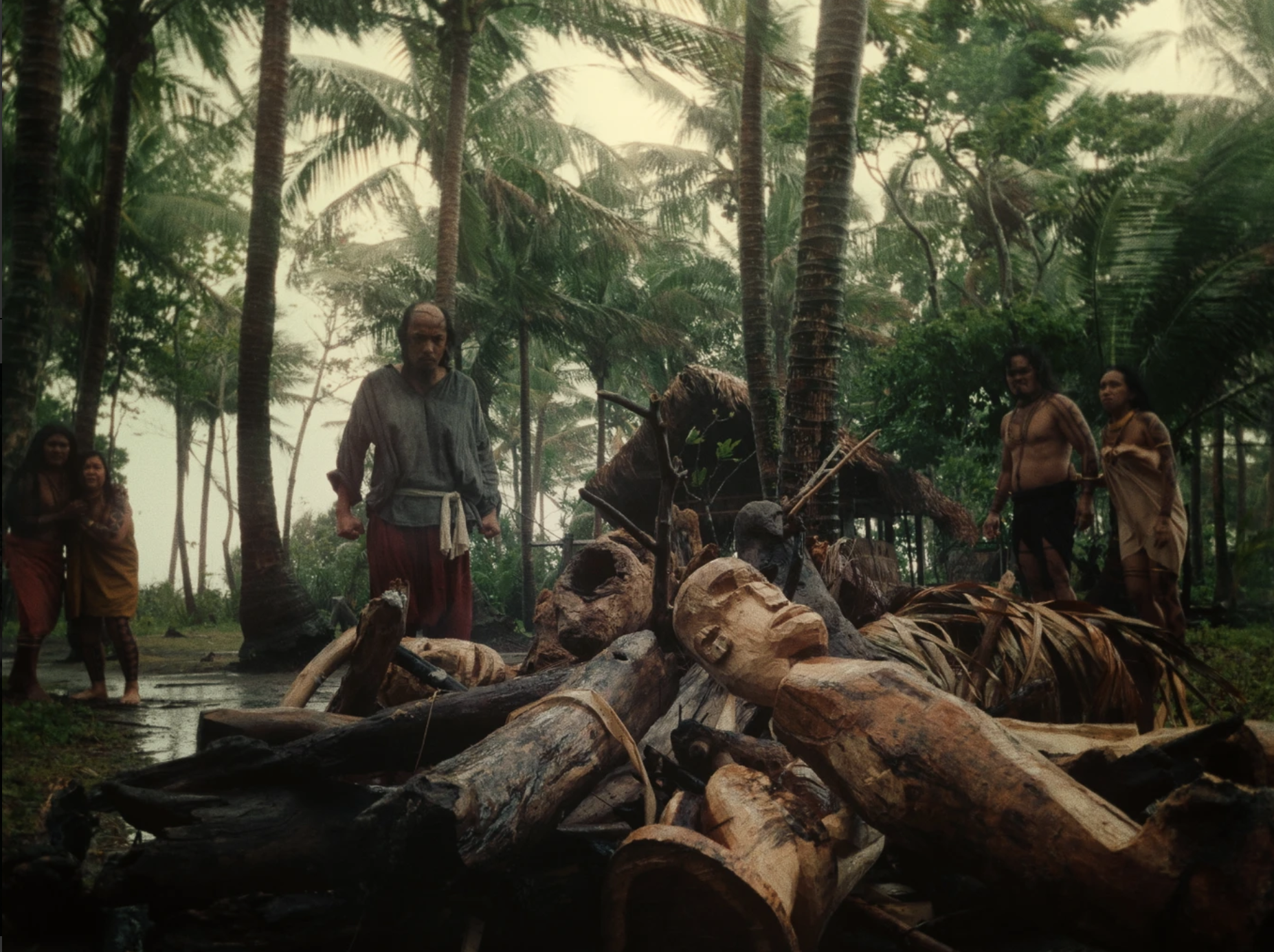 A man stands behind a pile of wooden idols visibly angered. Two distressed native couples stand on either side of the pyre. A small hut is visible in the background as well as a dense forest of palm trees.