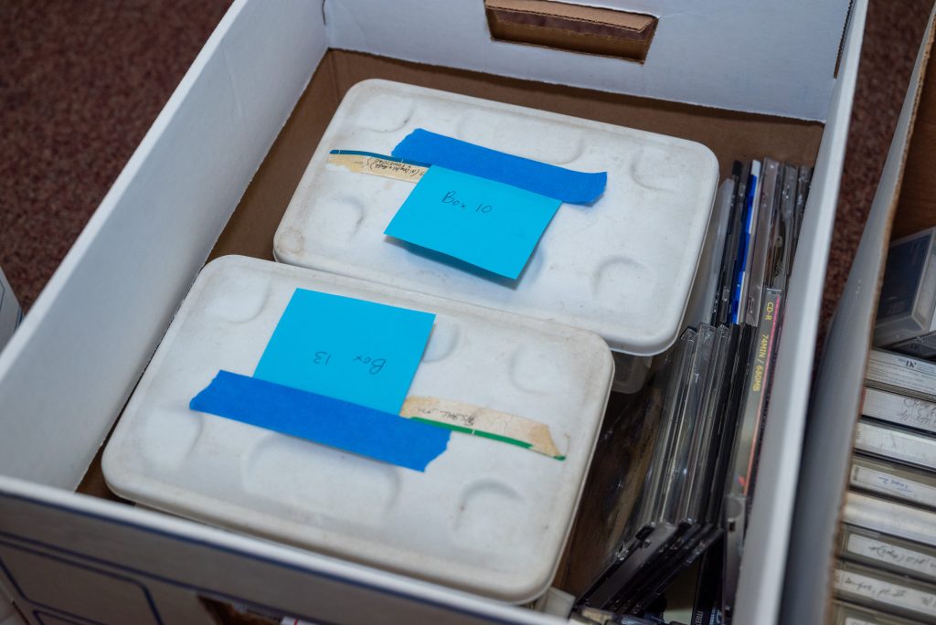 A cardboard box filled with HoneyPot Performance archive materials sits on a pinkish-brown carpet. Inside are two small plastic totes labeled ‘Box 13’ and ‘Box 10,’ written on a blue sticky note with blue tape. To the right in the same box are a couple of stacks of CDs. Image by Tonal Simmons.
