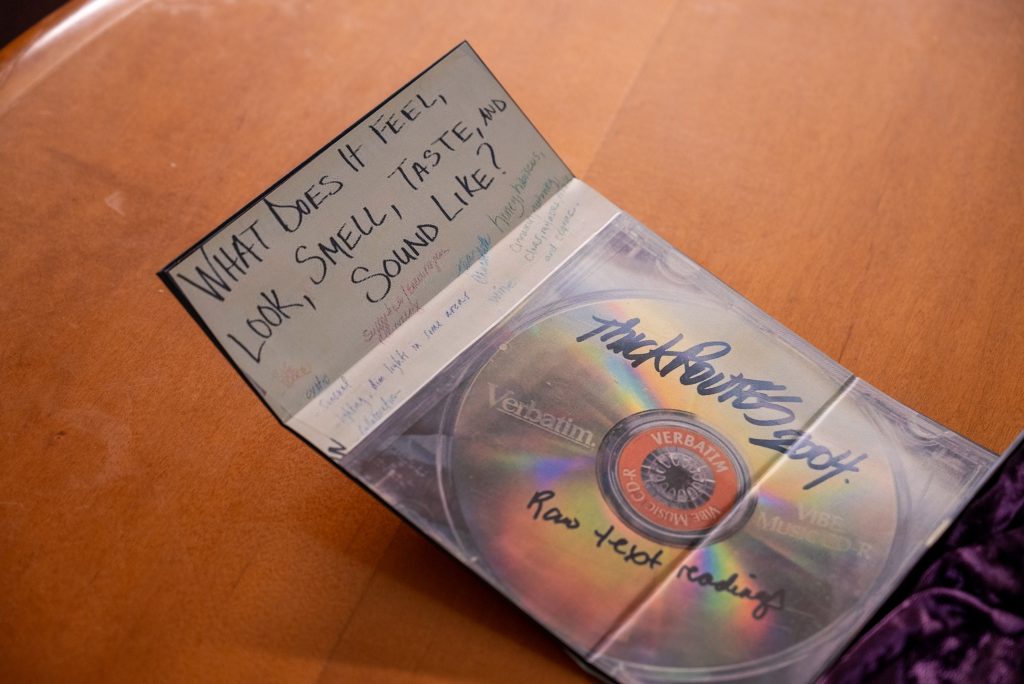 A partial shot of a handmade archive box created by Siobhan McKissick, made of items and images from HoneyPot Performance. The interior of the box contains two images, one of a physical CD with the following text handwritten: “Thick Routes 2004, Raw Text Readings.” The image to the left on the other flap has text written that says, “What does it feel, look, taste, and sound like?” It includes a list of handwritten responses from HoneyPot members in various colors. Image by Tonal Simmons.