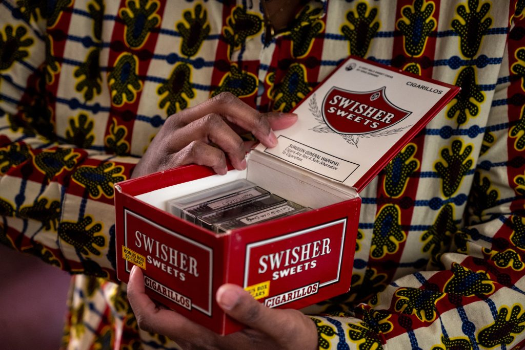 A close-up shot of Siobhan McKissick holding a recycled red Swisher Sweets cigar box repurposed to hold physical tapes of HoneyPot’s archived recordings. She holds with them two hands holding the box to expose the tapes. 
 Image by Tonal Simmons.