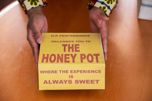 A close-up shot of the handmade archive box held between Siobhan’s hands as it sits on a brown table. The top of the box features a picture of an archived yellow flyer wrapped on the flap that reads in red capitalized text: “H.P. Performance Welcomes you to The Honey Pot Where the Experience is Always Sweet.” Photograph by Tonal Mondae.
