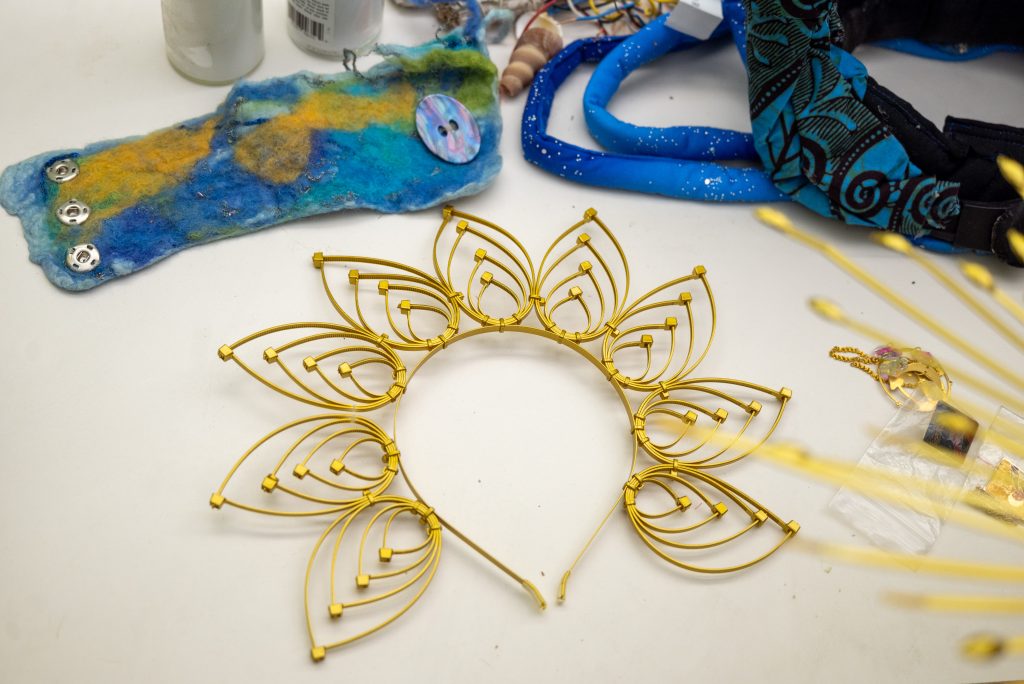 A crown made of small zip ties, wrapped around a headband featuring eight teardrop shapes spread out, painted gold, sits on the table surrounded by other Honey Pot ephemera. Image by Tonal Simmons.