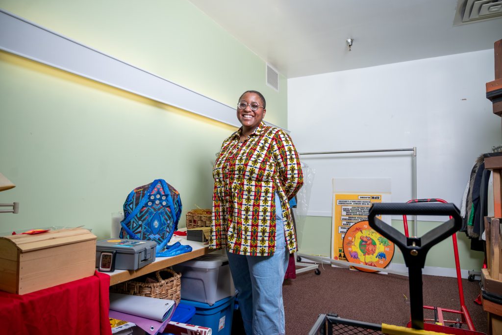 Siobhan McKissick stands with her arms behind her back in the middle of one of Honey Pot’s archival rooms as she looks directly at the camera with a smile. She is wearing blue jeans with a long sleeve African print shirt with yellow, red, black, blue, and cream. Honey Pot ephemra sits in the background. Image by Tonal Simmons.