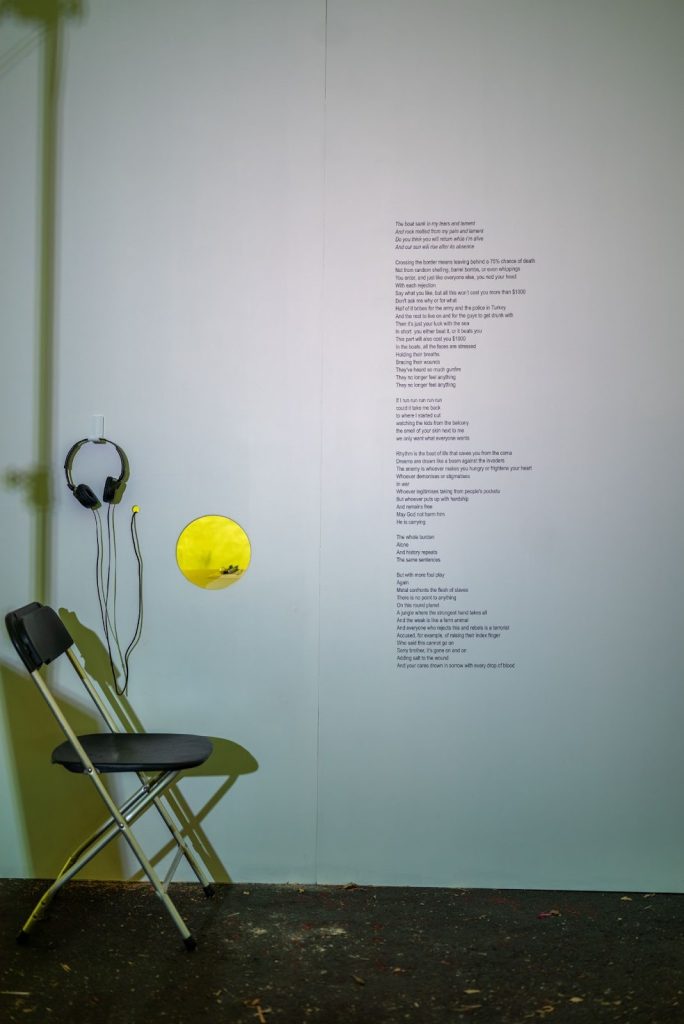 The As Far as My Fingertips Take Me artist booth features a white gallery wall with a circular hole cut into it. To the right, lyrics from a song by Basal Zarra are displayed directly on the wall. To the left, a pair of black headphones hangs with its cord fed back into the wall. Below the headphones sits a black-and-gray folding chair. Photo by Tonal Simmons.