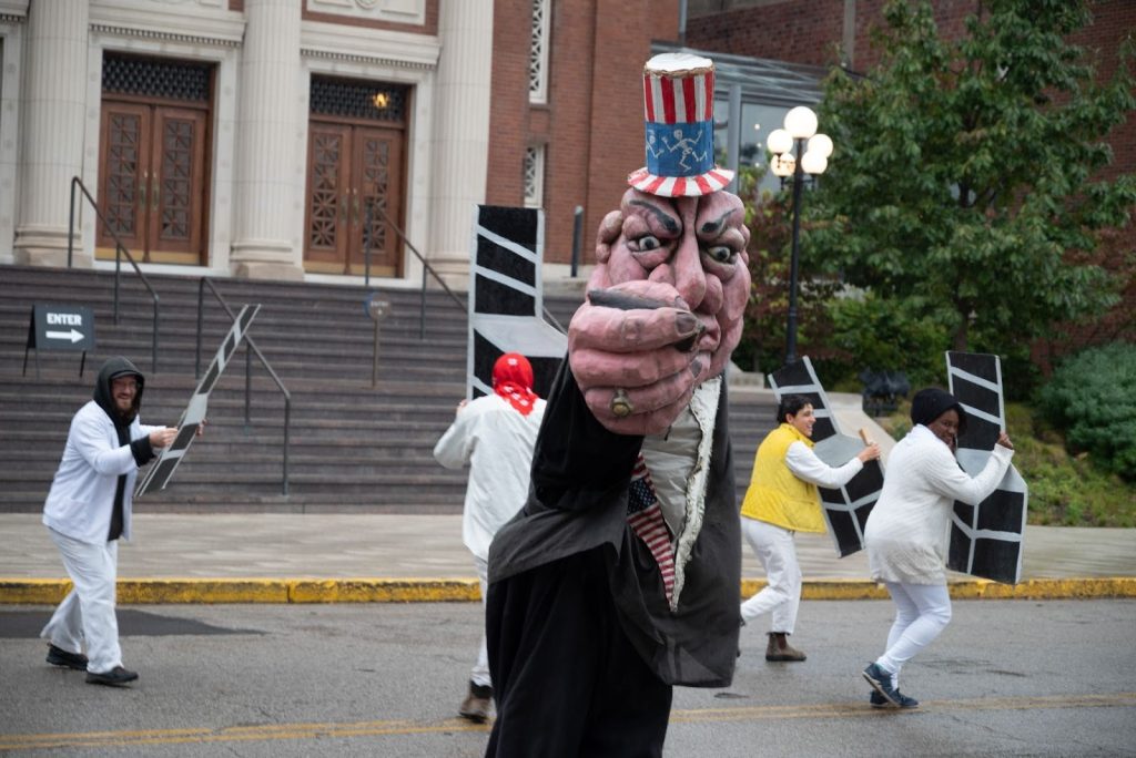 Bread and Puppet Theatre performs in the middle of a street while marching toward CounterPublic for a communal lunch. Four performers dressed mostly in white hold white chairs painted on black paper as they move around a large pink figure. The figure wears a black robe, holds a cigar, and wears a red, white, and blue top hat resembling those associated with politicians. The figure is faced towards the camera as it gets ready to swing it’s fist at the chairs. Photo by Tonal Simmons.