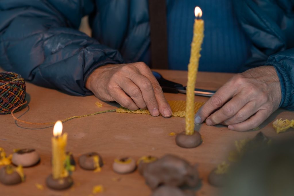 A person wearing a blue jacket rolls yellow beeswax onto a rainbow-colored wick at the Spellcasting Booth. In front of them, a candle rests inside a handmade clay candle holder on a table covered with brown parchment paper. Several other participant-made candle holders sit near the front of the image. Most are shaped like rocks, and one is shaped like a buffalo. Photo by Tonal Simmons.