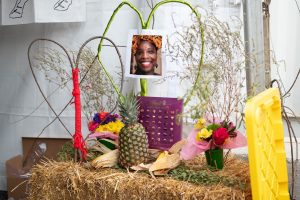 Image: An altar made of hay honors Patrisse Cullors, artist, activist, and co-founder of Black Lives Matter. She was unable to attend The Circus after her home was unlawfully raided by police. Created by her co-creators from the Spell Casting booth, the altar features two small, colorful flower bouquets in green vases placed at each end. Between them sit a pineapple, an ear of corn with its husk, and a purple box of Lindt chocolates. Two hearts made from sticks rest nearby—one wrapped in green yarn and the other in red. A printed photo of Patrisse Cullors is placed at the center of the green heart. Photo by Tonal Simmons.