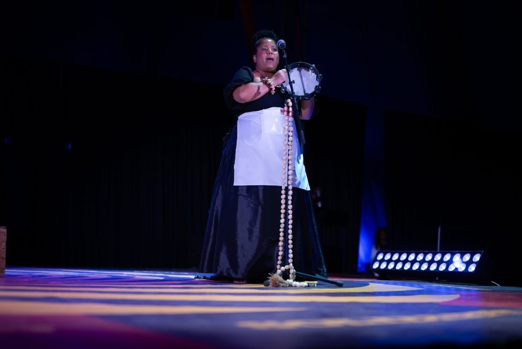 Rashida Bumbray stands upright, singing into a microphone while playing a tambourine. A large, light-brown beaded strand hangs from the microphone stand and extends to the stage floor. She wears an all-black dress with a white apron and brown beaded bracelets on each wrist. The stage floor is patterned in dark blue, red, and yellow in a circular design. Stage lights sit on the floor to her right, and black curtains form the background. Photo by Tonal Simmons.