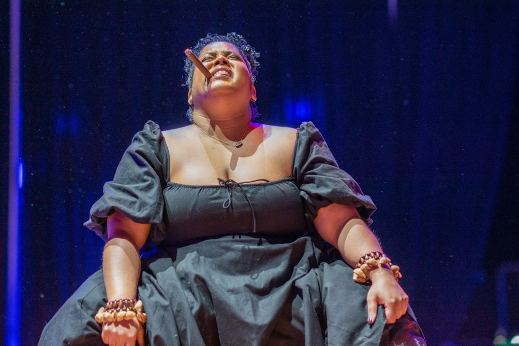 Rashida Bumbray sits upright with her face tilted upward and her eyes closed. A stage cigar is held between her teeth on the right side of her mouth. Her hands grip the black fabric of her dress at knee level. She wears brown beaded bracelets on both wrists, and her hair is pulled back into a high ponytail, with curls extending toward the back curtain. Photo by Tonal Simmons. 