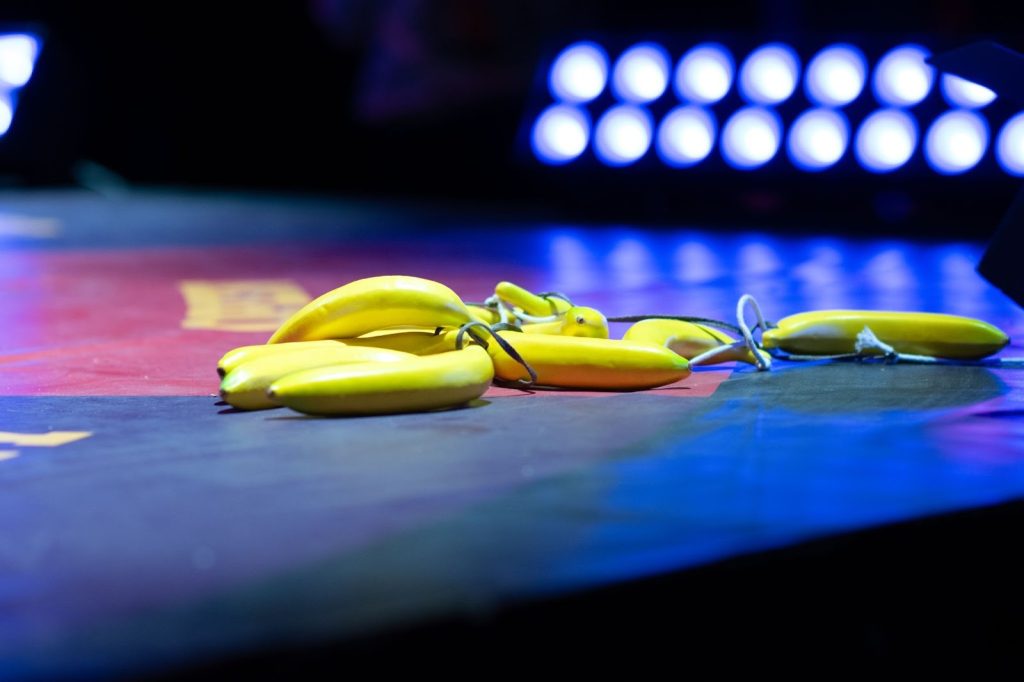 A cluster of yellow plastic bananas from the performer’s bustle lies across the stage floor, tied together with black and white string. The floor is patterned in dark blue, yellow, and red. Stage lights sit on the floor in the background. Photo by Tonal Simmons.