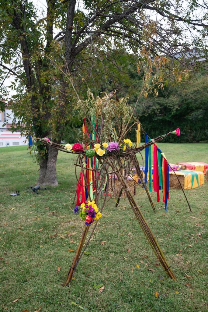 A bison sculpture stands about four feet tall, constructed from multiple sticks shaped into four legs and a body. Colorful flowers and ribbons fill parts of the face and horn area. Hay benches from The Circus sit nearby, and a large tree rises in the background. Photo by Tonal Simmons.