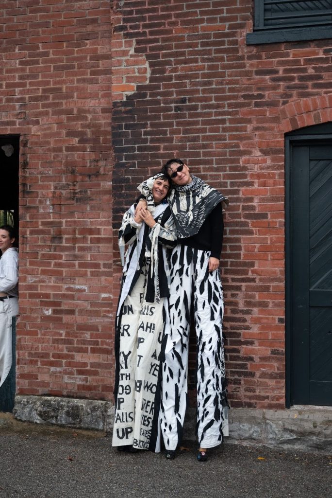 Two members of Bread and Puppet Theatre stand on stilts, leaning back against the red brick exterior of CounterPublic. They hold onto each other side by side and smile toward the camera. Both wear black long-sleeve shirts and patterned black-and-white pants. Photo by Tonal Simmons.