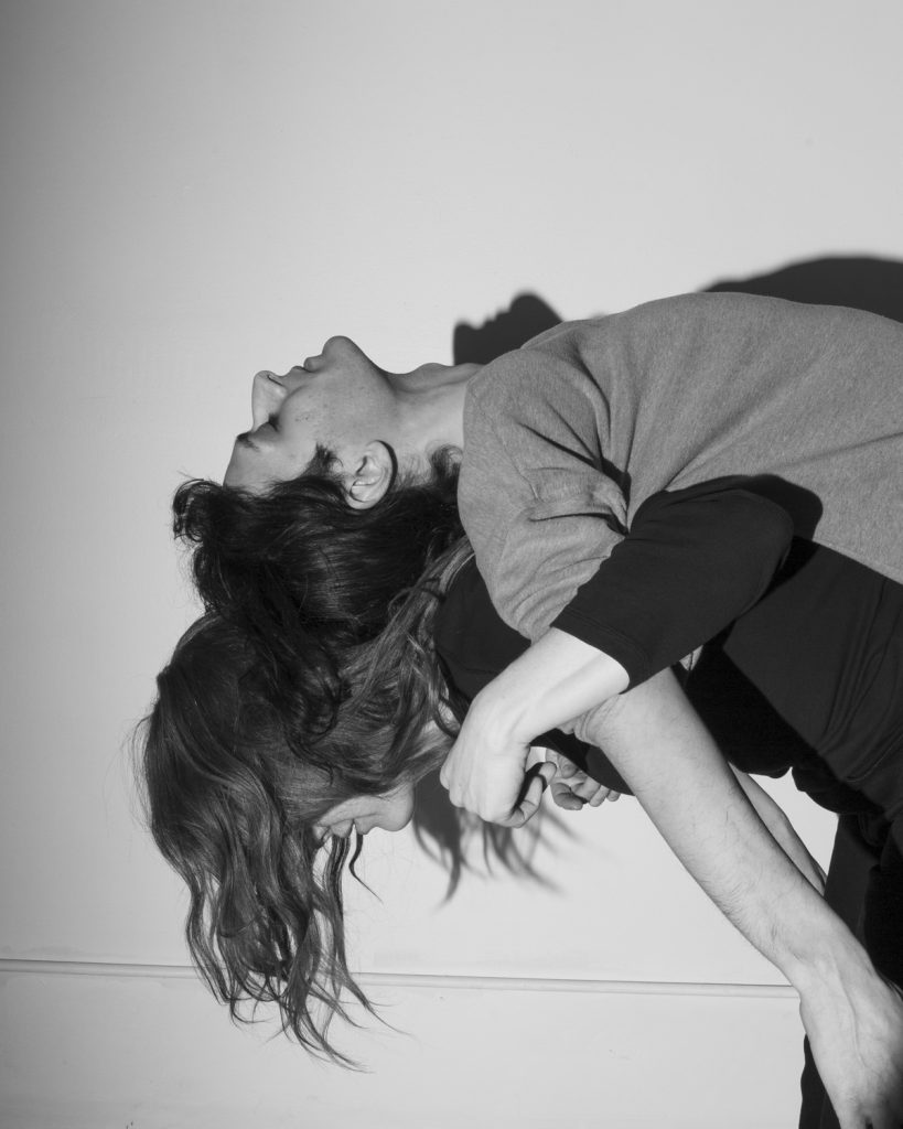 A photograph titled "Untitled (Being Hesam’s Mother)" from the series Mother Figure by Kat Bawden, 2025. A black and white photograph of a woman carrying a young man on her back, their arms interlocked. She bends over forward, and her hair covers her eyes. He rests upon her on his back, eyes closed, with a calm facial expression. 