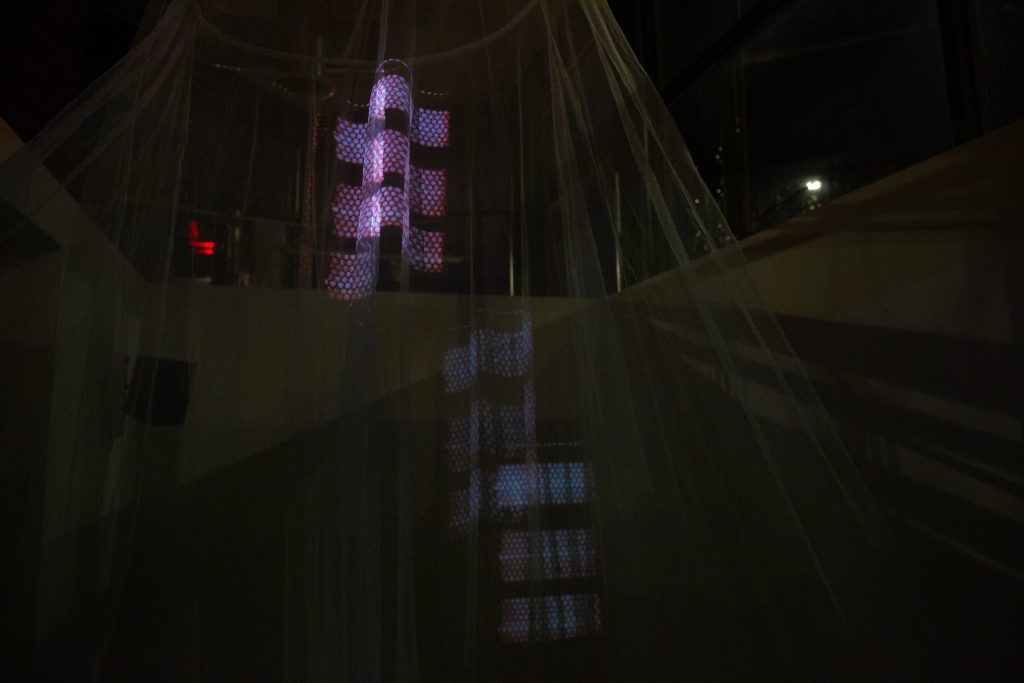 A close-up photograph of an installation shows projected images of a radiator on a suspended mosquito net and on a wall behind it in a dark room. That one time I felt loved when I was little by Ruby Que Digital projection, sound (lullaby), mosquito net,  2025, photograph by Ruby Que. 