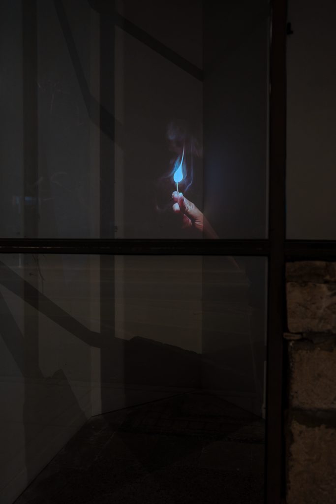 A projected image shows a hand holding a burning match in a dark space. Little Match Girl by Ruby Que Video projection (viewed from the storefront window of Roots & Culture), 2025, photograph by Ruby Que. 