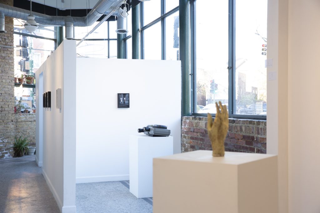 A general image of the gallery space that shows as follows from the left: a wall partition with small photographs in black frames in the background, a carousel projector on a pedestal facing one of the walls in the middle ground, and a golden hand on a pedestal in the foreground. Right to left: Interactive carousel projector for Kat Bawden’s photographs and Sau Zuk by Ruby Que, cast pewter, 8.5” x 4” x 2”,  2025, photograph by Kat Bawden. 