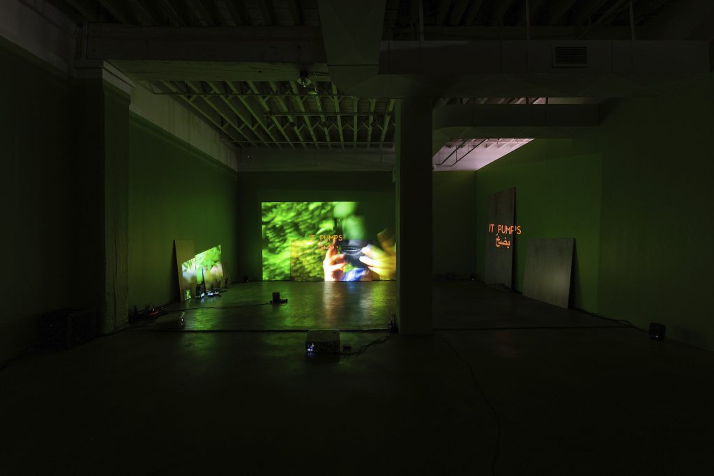 A three-channel video projection features Arabic script, the phrase "IT PUMPS" and hands holding a Neolithic mask against a green background. There are several panels leaning against the walls of the gallery and a few speakers and projectors arranged throughout the room. Photo by Colin Conces. 
