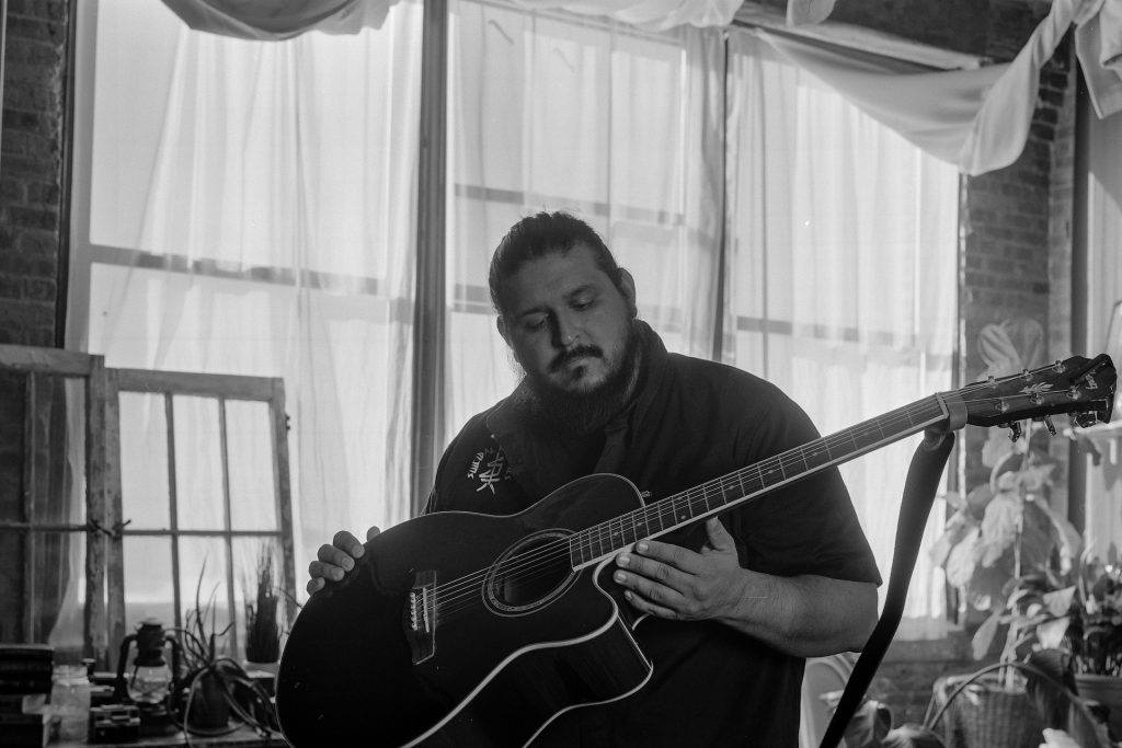 Black and white captures of photographer Carlos Martinez sitting in his studio and holding his guitar. Photos by Genesis Falls.
