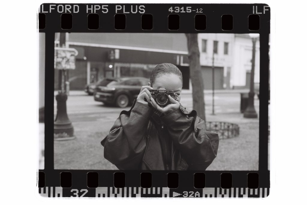 A black and white film scan captures photographer Han on cam holding her camera to her face as if she is taking a photo simultaneously. Photo by Genesis Falls.