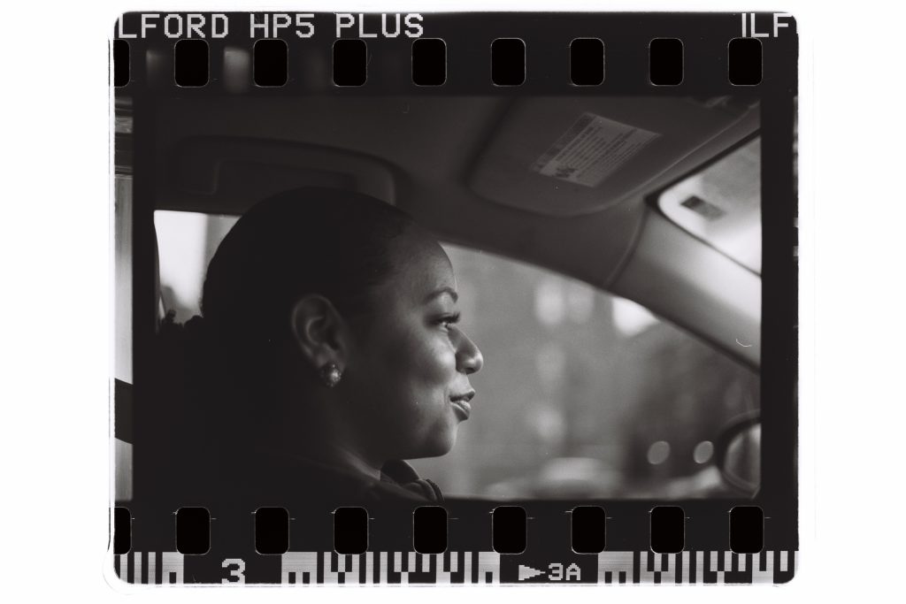 A black and white film scan captures a side view of photographer Han on cam sitting in the driver's seat of a car. Photo by Genesis Falls.