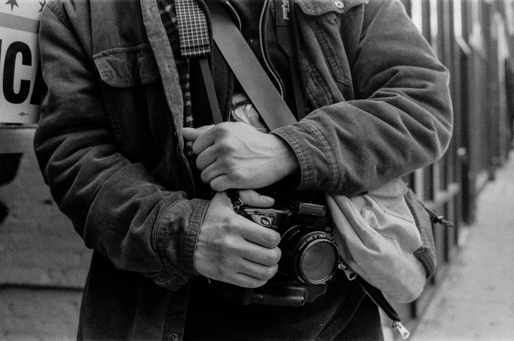 A black and white film capture of photographer Josue Cuyun's torso in the frame holding his camera that is hanging on his neck. Photo by Genesis Falls.