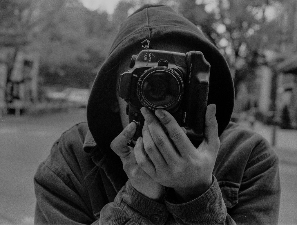 A black and white film capture of photographer Josue Cuyun holding his camera to his face as if taking a photo simultaneously. Photo by Genesis Falls.