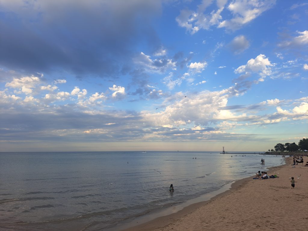 A landscape photo of Foster Beach. Some people sit on the sandy beach, while others tread in the calm, blue water. The sky fades from a vibrant blue into a pale yellow at the horizon. White clouds dot the sky. Photo courtesy of Emily McClanathan.