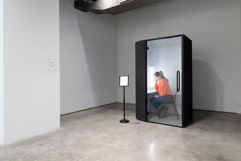 A woman sits inside a soundproof booth in a gallery space. The door of the booth is transparent. The woman is wearing jeans and an orange sweater. There is a sign positioned beside the booth. Photo by Colin Conces. 