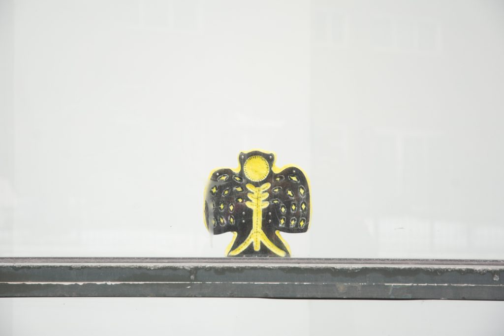 Sending a Picture (owl), 2026. A yellow and black owl sculpture made from fresco and wood is displayed on a windowsill facing the street. There is an abstract pattern on the owl's body and stomach. Its face is a blank yellow circle.