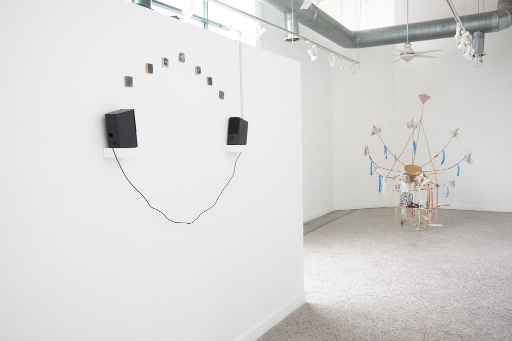 Two small speakers are mounted on wall shelves facing each other with a wire dangling between them. Seven ceramic tiles with geometric patterns on them are affixed to the wall in an arc between the speakers. There is an assemblage sculpture that resembles a satellite transmitter in the central gallery space. 