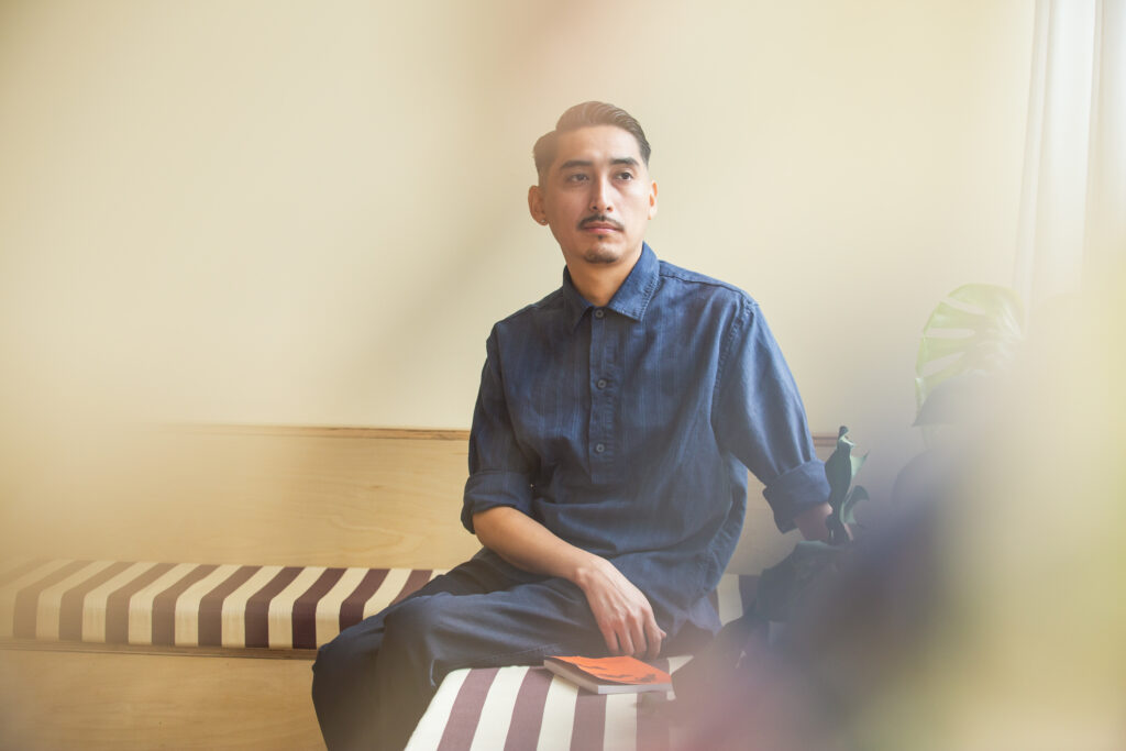 John H. Guevara sits on a bench in the front room at Tala Gallery with a red book at his side. He is wearing an all blue outfit and is looking away from the camera with a smirk on his face. Image by Kristie Kahns.