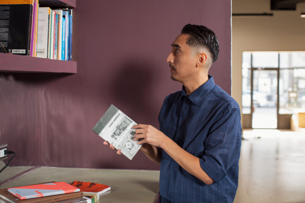 John H. Guevara holds a grey colored book while looking at a book shelf at Tala Gallery. He is wearing a blue button up top. Image by Kristie Kahns.