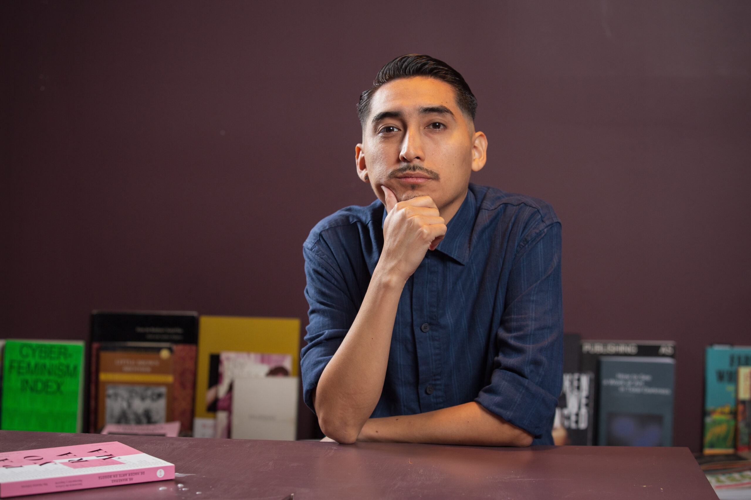 John H. Guevara sits on a table in the back room at Tala Gallery with books in front of him. He is looking straight at the camera with a smirk on his face. Image by Kristie Kahns.