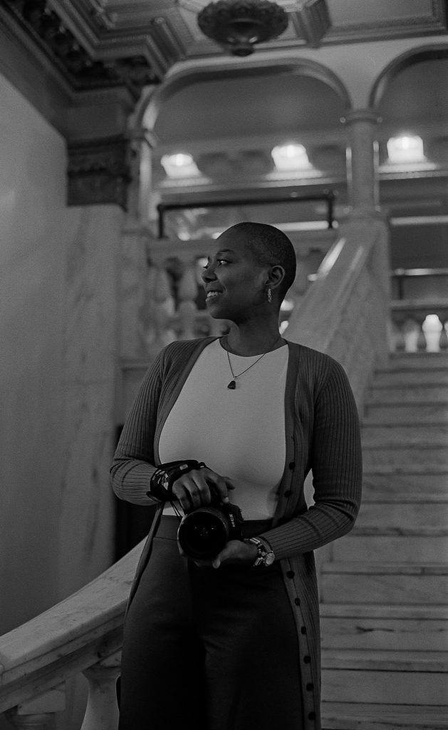 Black and white captures of photographer Sam Wilson holding her camera in her hands while smiling and standing at the bottom of a white staircase. Photo by Genesis Falls.
