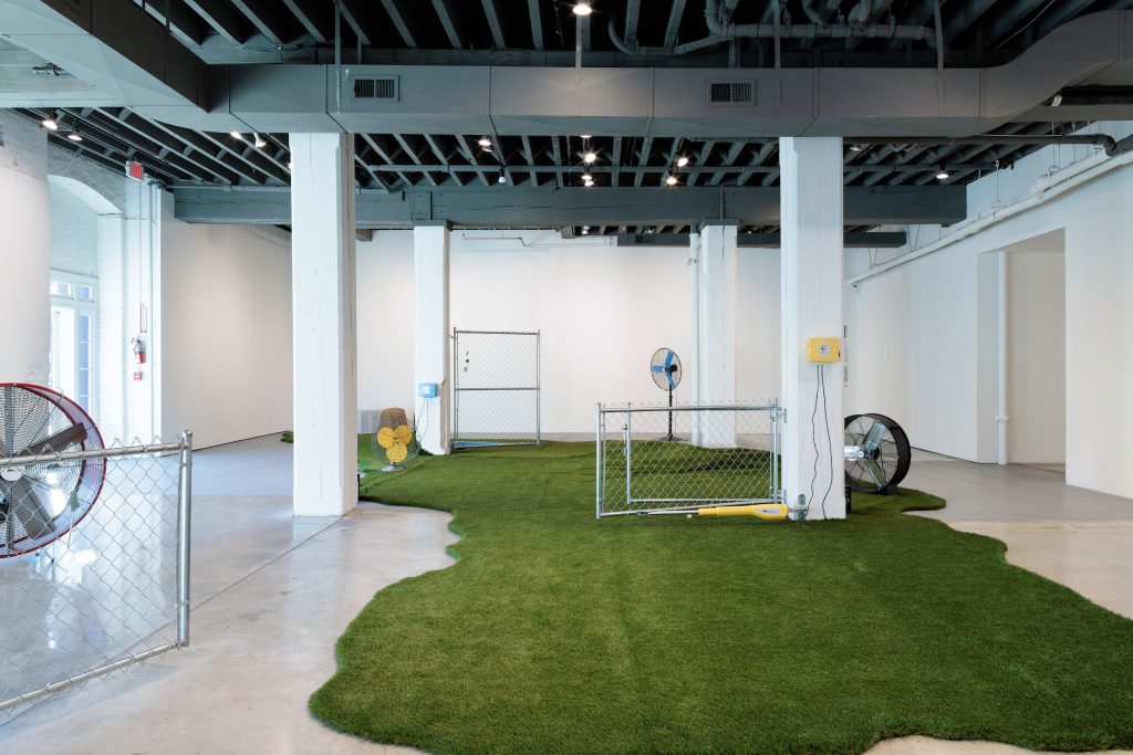 Artificial turf fills the center of a gallery space flanked by four concrete pillars. There are two large floor fans, one positioned on the turf and another on the gallery floor. There are two smaller fans on the far edge of the turf and two chainlink fences attached to columns on the left and right side of the room and another fence in the foreground. There are several speakers positioned near the columns as well. Photo by Colin Conces. 