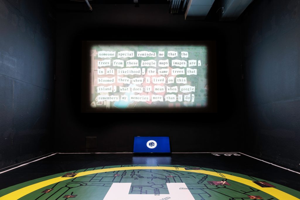 An image of cut-up text is projected onto a blackbox theater wall. There is a monitor underneath the projection with the image of a cursor against a black background. The floor has a circular pattern of green and yellow with lines that resemble circuitry. Photo by Colin Conces. 
