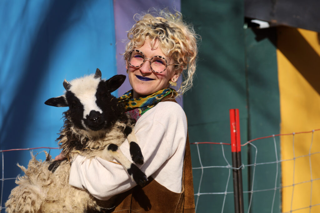 Rachel smiling in the pasture while holding one of their black and white, 2025-born lambs, Disco. Rachel is seen wearing round tortoiseshell glasses, dark purple lipstick, and a loose long-sleeved white top. A colorful striped tarp hangs in the background. Image by Viki Stark.