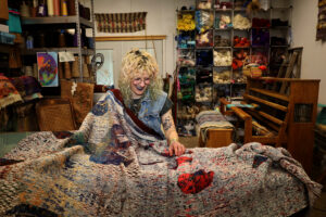 Rachel sitting in the studio while showing off her piece “[Saccharomyces]”, the largest of the pellicle weavings. The piece includes cool-toned reds and blues, as well as yellows, teals, and creams and is a 83” x 16’8” handwoven Jacquard. The pellicle weavings use blending of color and patterns and shapes to illustrate the way yeast (or communities of yeast, i.e. pellicles) recycle physical materials in our world. They are flat and sometimes intentionally confusing, textural abstractions. To the right is the yarn shelving wall and their 16 Shaft Leclerc Loom. To the left are additional supplies resting on a shelving unit, filled with cones, skeins, and more. Photo by Viki Stark.