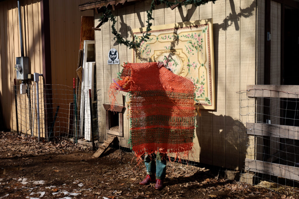 Rachel stands in the sun while in Puzzle the donkey’s pasture, holding the baling twine piece over her head. A floral painting hangs behind her, affixed to the chicken coop’s outer wall. Image by Viki Stark.