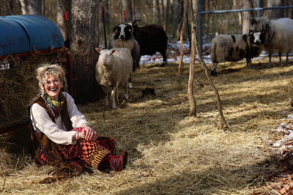 Rachel sitting and smiling in the pasture with lambs in the background. Image by Viki Stark.
