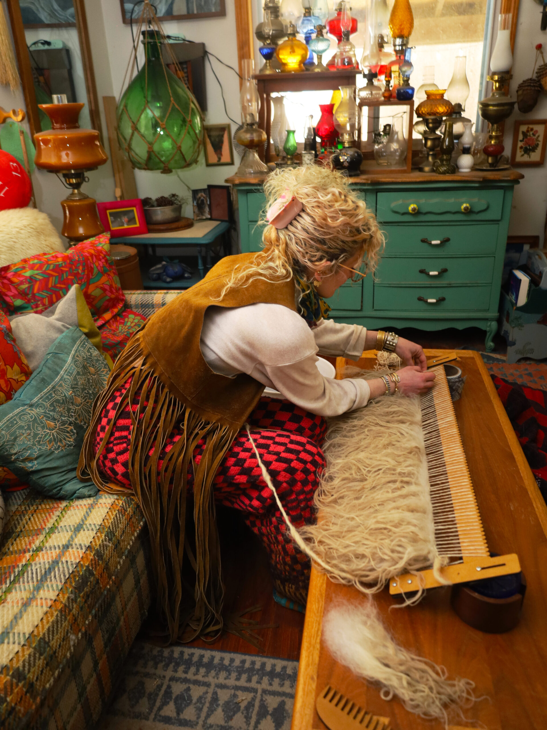 Rachel sits on a plaid couch in her home while wearing self-made red wool-checkered pants, a brown fringed vest, and her blonde curls are up in a claw clip. She leans over as she weaves on the Varafeldur piece. The home has a wide assortment of colorful glass lamps on a teal dresser in the background and an arrangement of framed art on the walls. Image by Viki Stark.