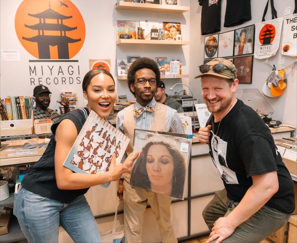 Virginia and two other people are smiling for the camera with their vinyl at Miyagi Records. Shelves with mugs, posters, books, records, tote bags, and other merchandise are displayed at the checkout spot behind them. Photograph by Wendy Davis.