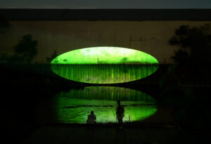 Xingyu Huang, Beneath Breathing, 2025. Site-specific installation, Chicago River, IL. 2:30 min. Documentation photography. Photo by Yousif Alzayed. Courtesy of the artist. The image captures a large green oval-shaped video projection glowing on the façade of a dark riverside warehouse at night, mirrored in the water below. Two people stand and crouch on the riverwalk in silhouette, watching the luminous reflection spread across the river’s surface.