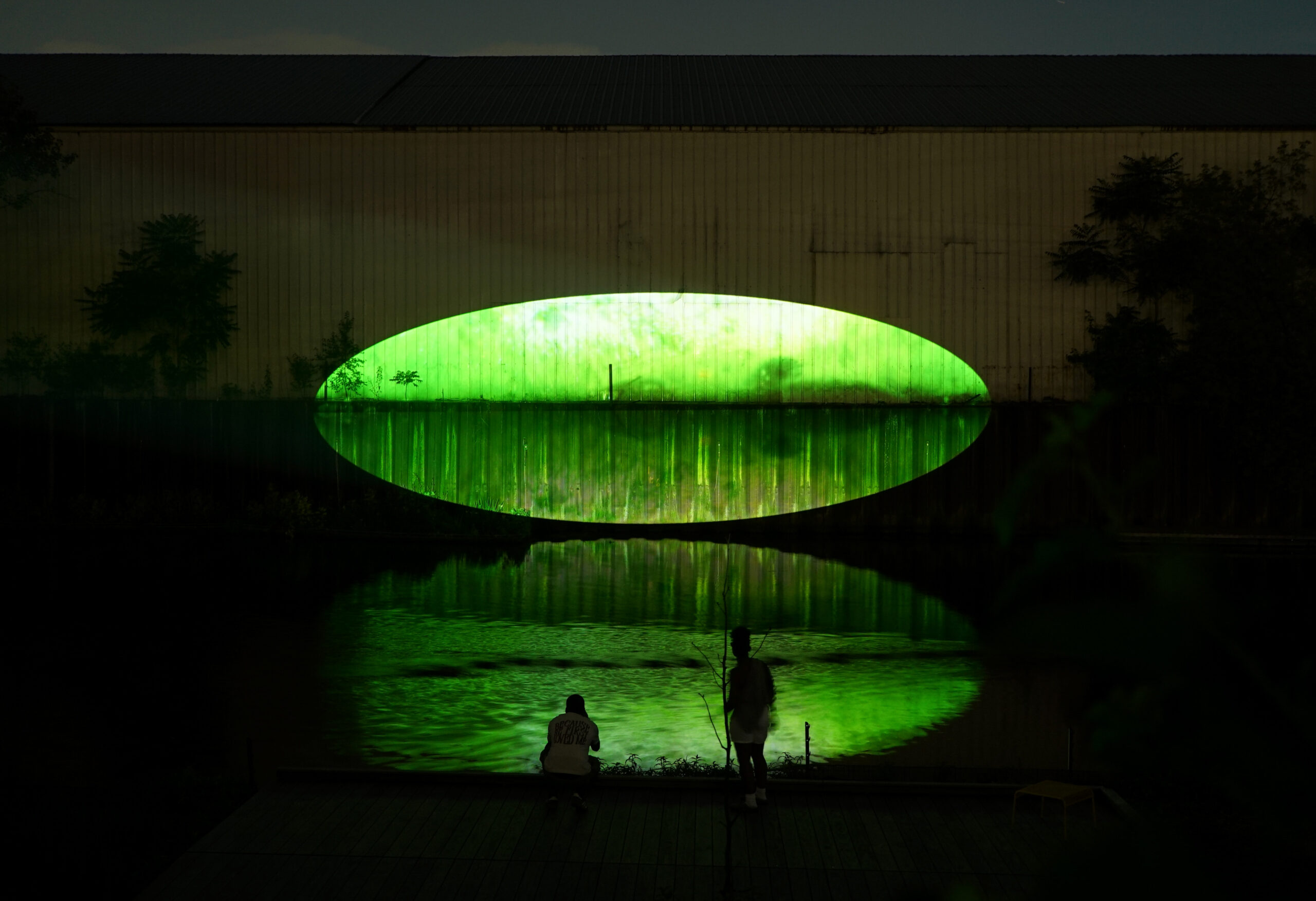 Xingyu Huang, Beneath Breathing, 2025. Site-specific installation, Chicago River, IL. 2:30 min. Documentation photography. Photo by Yousif Alzayed. Courtesy of the artist. The image captures a large green oval-shaped video projection glowing on the façade of a dark riverside warehouse at night, mirrored in the water below. Two people stand and crouch on the riverwalk in silhouette, watching the luminous reflection spread across the river’s surface.
