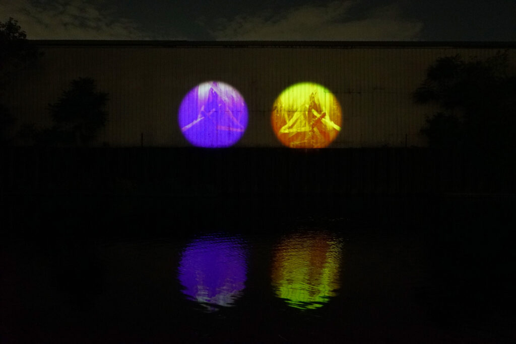 Xingyu Huang, Beneath Breathing, 2025. Site-specific installation, Chicago River, IL. 2:30 min. Documentation photography. Here are two circular projections, one purple and one yellow-green, cast fisheye-camera views of a muddy riverbed and underwater plants onto the wall of a dark warehouse beside the river at night. The color contrast, with purple standing in for ultraviolet, uses the fisheye perspective to suggest a mode of seeing beyond human vision and closer to the expanded visual spectrum available to many fish. Photography by Yousif Alzayed courtesy of Xingyu Huang.