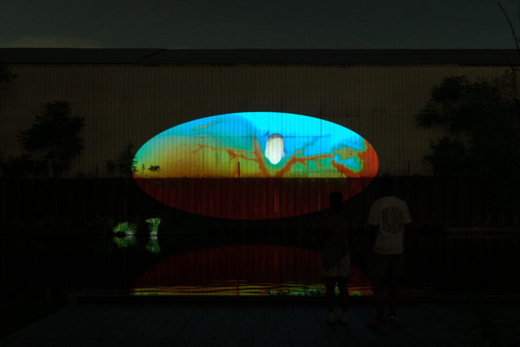 Xingyu Huang, Beneath Breathing, 2025. Site-specific installation, Chicago River, IL. 2:30 min. Documentation photography. The image shows a projection of reversed underwater and above-water footage on the wall of a dark warehouse beside the river at night. The blue and orange moving image shows underwater plants and fish, while in the lower left corner, reversed human footsteps on the riverwalk appear as a small glowing reflection. Photography by Yousif Alzayed courtesy of Xingyu Huang.