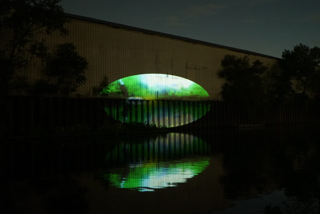 Xingyu Huang, Beneath Breathing, 2025. Site-specific installation, Chicago River, IL. 2:30 min. The image shows the same content as the first image: a large green oval-shaped video projection glowing on the façade of a dark riverside warehouse at night, mirrored in the water below. It is taken from a slightly tilted 45-degree angle from the left side of the projection. Photography by Yousif Alzayed courtesy of Xingyu Huang.