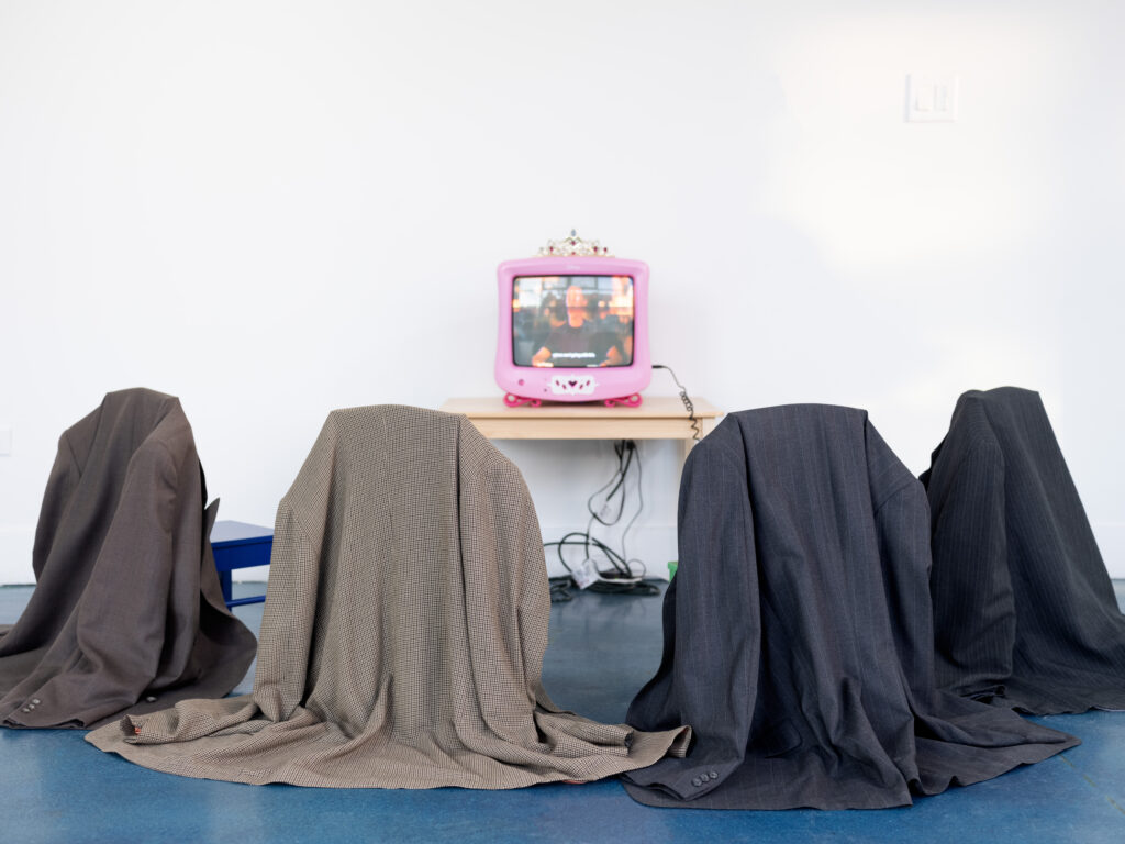 Installation view of Magifeifei's artwork Suck It Up! Buttercup!, a tiara-clad vintage Pink Disney CRT television, plays a scene from HBO’s The White Lotus. Four chairs surround the CRT in a semi-circle. Large blazers hang, draped on the back of each chair. Image courtesy of the artist. 