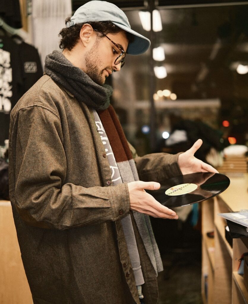 A person wearing a baseball hat, scarf, and light jacket looks closely at a record outside of its sleeve. Photograph by Austin Scott.